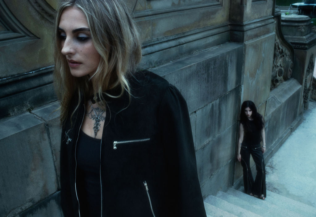 Woman in black jacket standing against a stone wall with another person in the background.