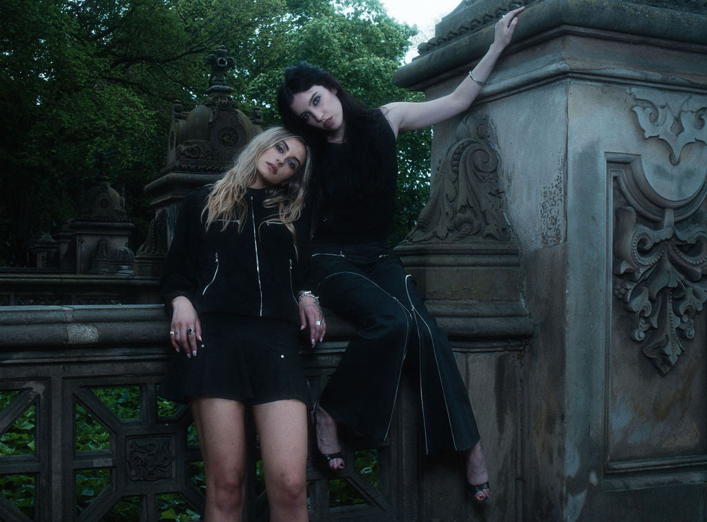 Two women in black outfits posing on a stone bench with a gothic background