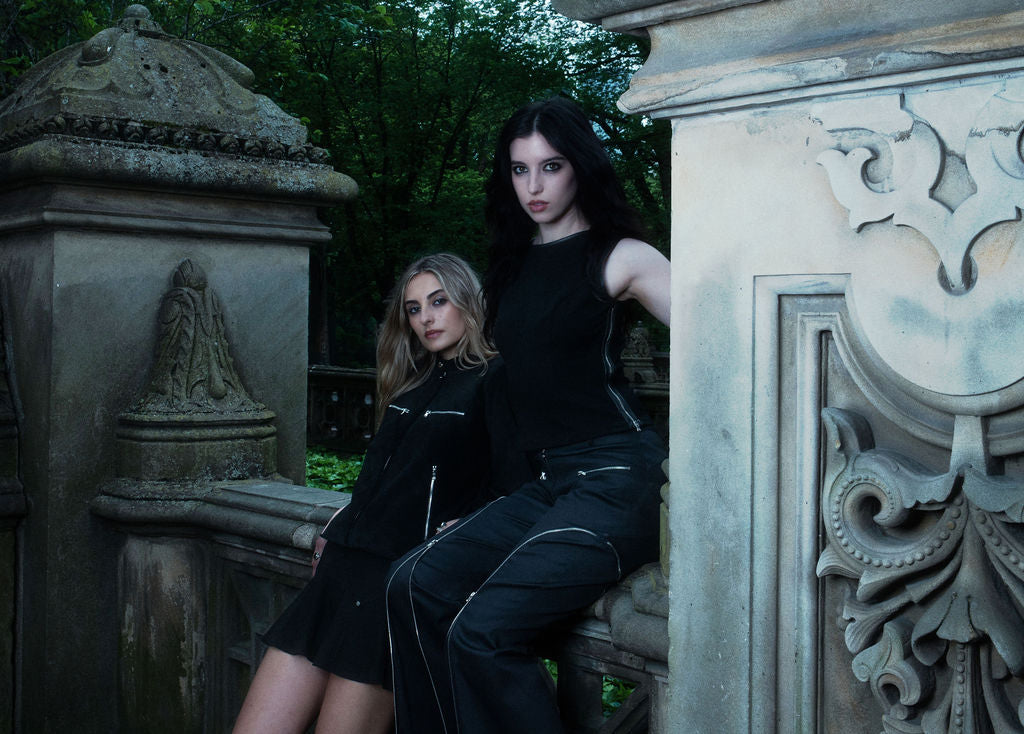 Two women in black outfits sitting against an ornate stone wall.