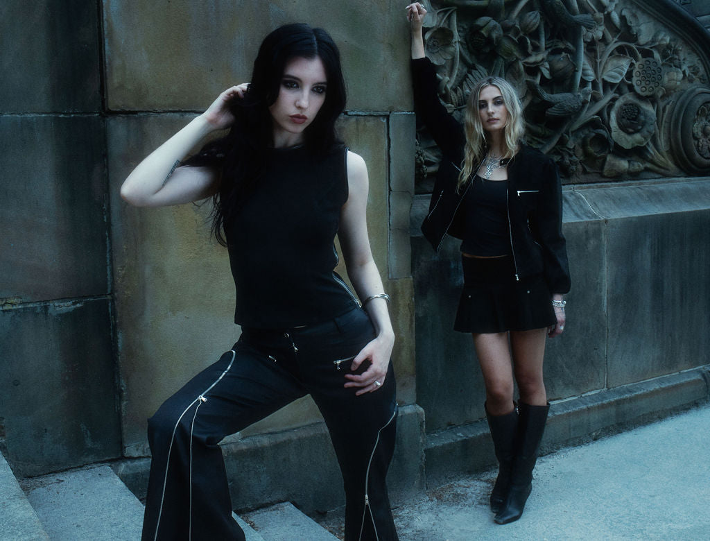 Two women in black outfits posing against a stone wall.