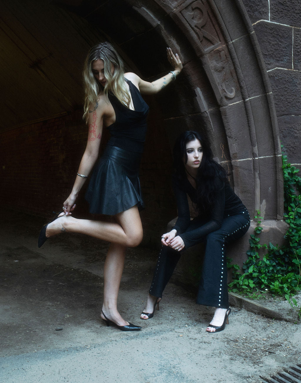 Two women in black outfits posing dramatically against a stone archway.