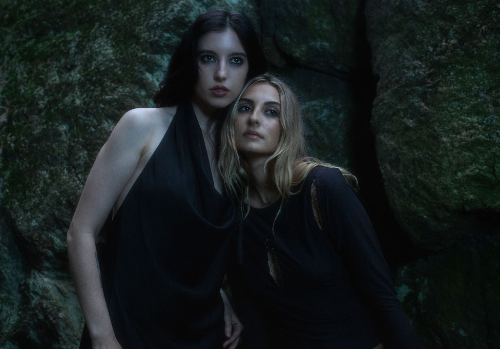 Two women in dark clothing standing against a textured wall.