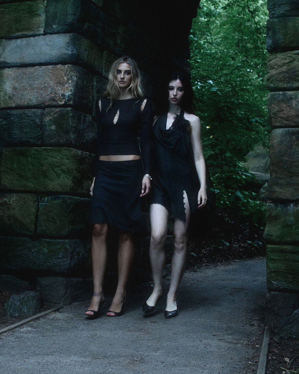 Two women in black outfits standing in a stone archway with greenery.
