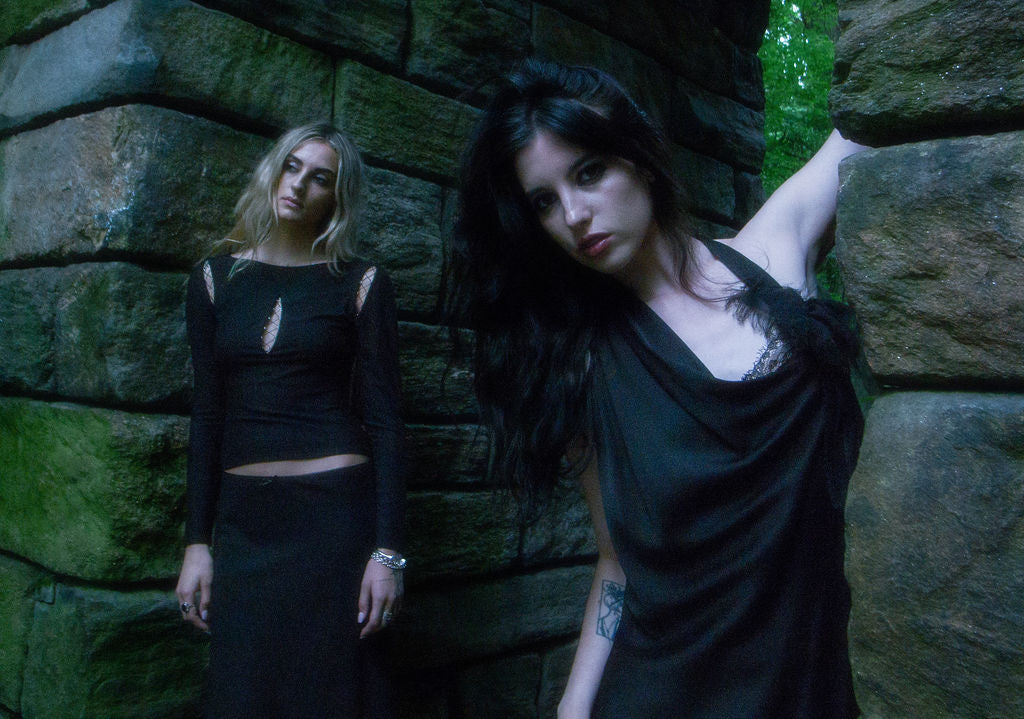 Two women in dark clothing standing against a stone wall with greenery.