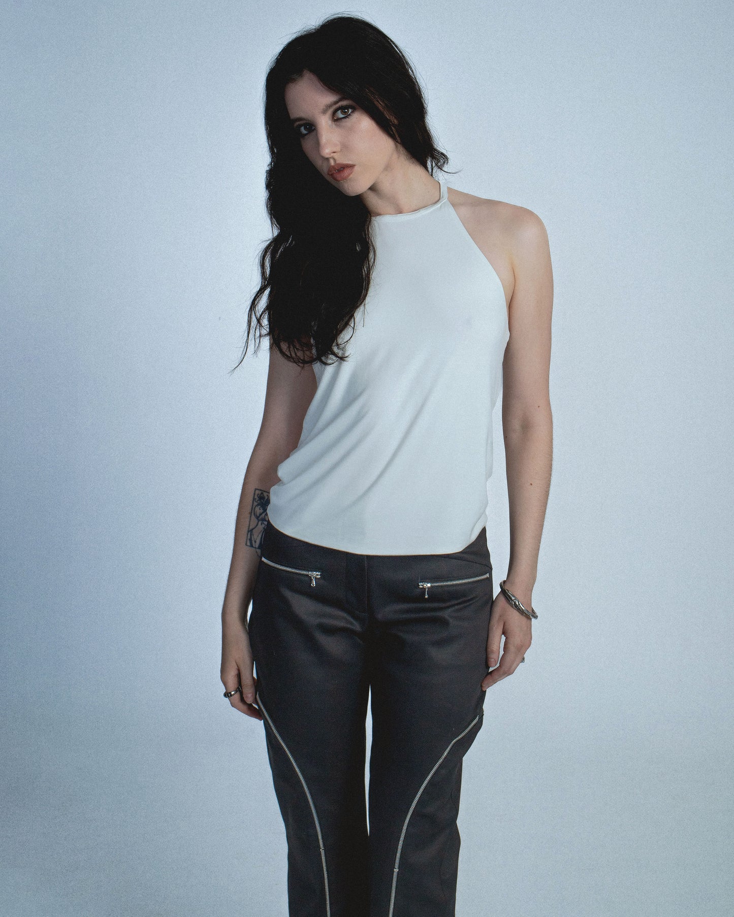 Halter neck top with a soft, fluid drape and open back. Twisted at the lower back with delicate strap detailing. Secured with an engraved silver studio Sivan logo clasp at the neck that clicks to open. Available in black and white. Black top features gunmetal clasp. Perfect for elevated day-to-night looks. Shop now at Sivan Studio.