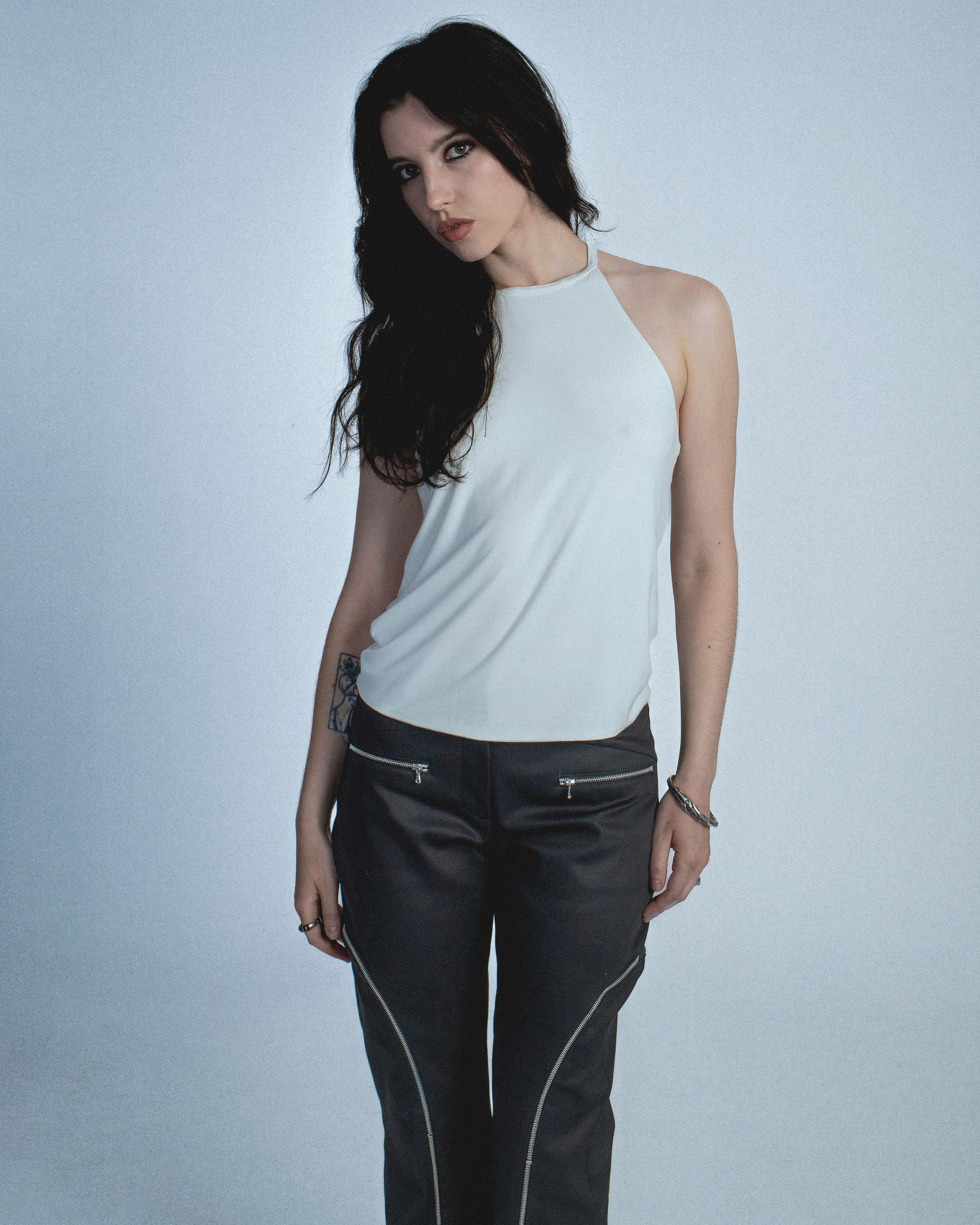 Halter neck top with a soft, fluid drape and open back. Twisted at the lower back with delicate strap detailing. Secured with an engraved silver studio Sivan logo clasp at the neck that clicks to open. Available in black and white. Black top features gunmetal clasp. Perfect for elevated day-to-night looks. Shop now at Sivan Studio.