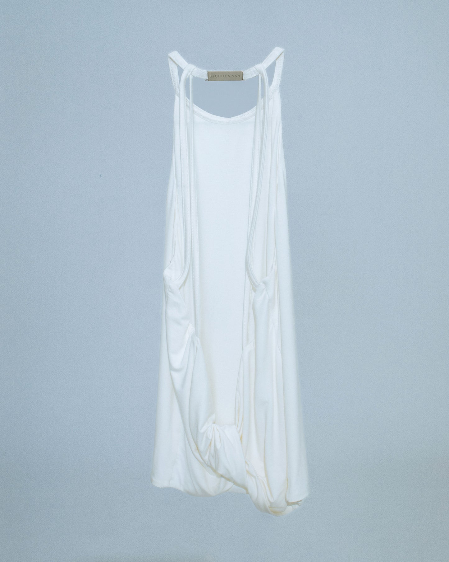 Halter neck top with a soft, fluid drape and open back. Twisted at the lower back with delicate strap detailing. Secured with an engraved silver studio Sivan logo clasp at the neck that clicks to open. Available in black and white. Black top features gunmetal clasp. Perfect for elevated day-to-night looks. Shop now at Sivan Studio.