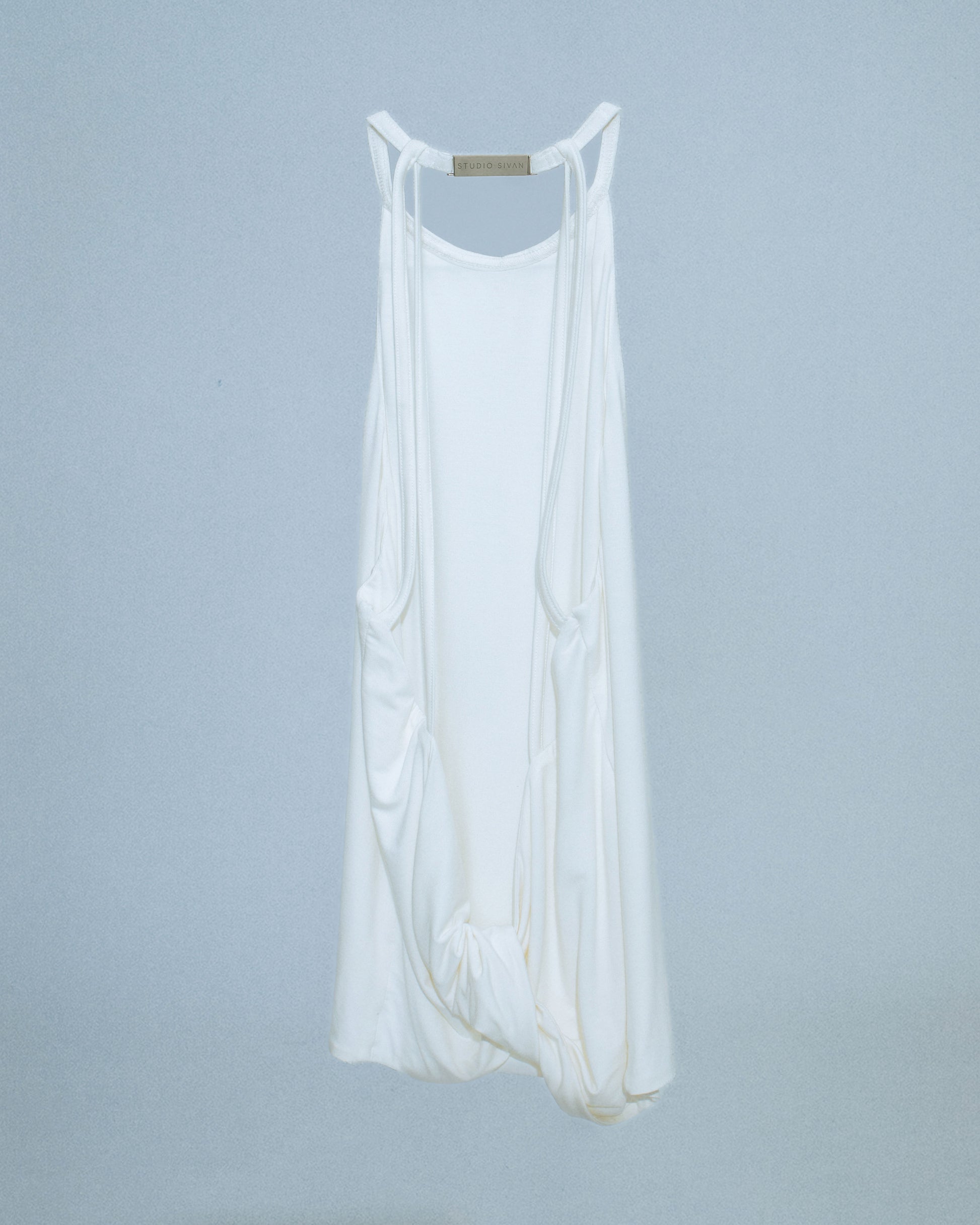 Halter neck top with a soft, fluid drape and open back. Twisted at the lower back with delicate strap detailing. Secured with an engraved silver studio Sivan logo clasp at the neck that clicks to open. Available in black and white. Black top features gunmetal clasp. Perfect for elevated day-to-night looks. Shop now at Sivan Studio.