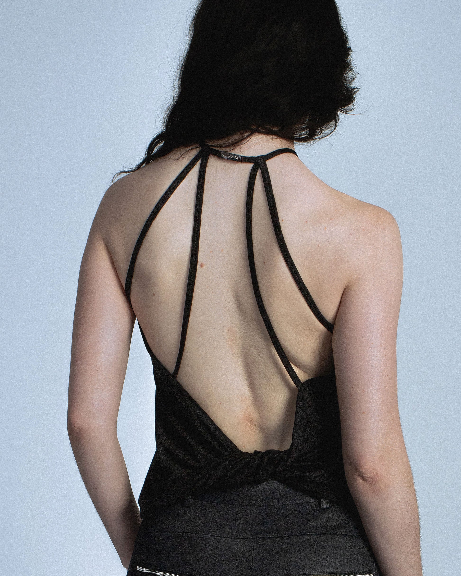 Halter neck top with a soft, fluid drape and open back. Twisted at the lower back with delicate strap detailing. Secured with an engraved gunmetal logo clasp at the neck. Available in black and white. Black top features gunmetal clasp. Perfect for elevated day-to-night looks. Shop now at Sivan Studio.